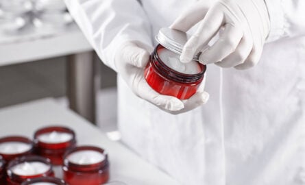 Um close-up elegante das mãos de um farmacêutico (com luvas, em ambiente limpo) manipulando ingredientes em uma balança de precisão digital ou misturando componentes em um béquer pequeno. O foco estaria nas mãos e nos elementos de manipulação, com o fundo levemente desfocado (um balcão de laboratório moderno e limpo, talvez com alguns frascos genéricos organizados)