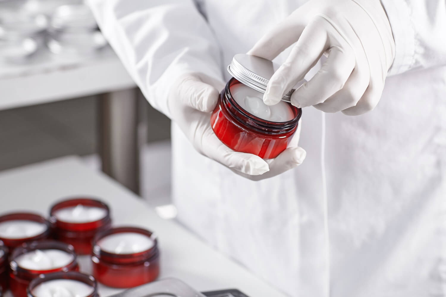 Um close-up elegante das mãos de um farmacêutico (com luvas, em ambiente limpo) manipulando ingredientes em uma balança de precisão digital ou misturando componentes em um béquer pequeno. O foco estaria nas mãos e nos elementos de manipulação, com o fundo levemente desfocado (um balcão de laboratório moderno e limpo, talvez com alguns frascos genéricos organizados)