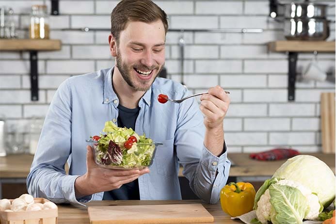 homem feliz e comendo um prato de salada na cozinha de casa