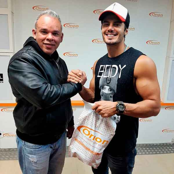 Adriano Garrido e Mariano Junior, um atleta ou influenciador fitness, apertando as mãos em um evento ou parceria com a Órion Farmácia de Manipulação. Mariano Junior segura uma sacola da Órion com o slogan 'Orion cuida de você', simbolizando a conexão da farmácia com o universo fitness, suplementação e a busca por saúde e bem-estar.