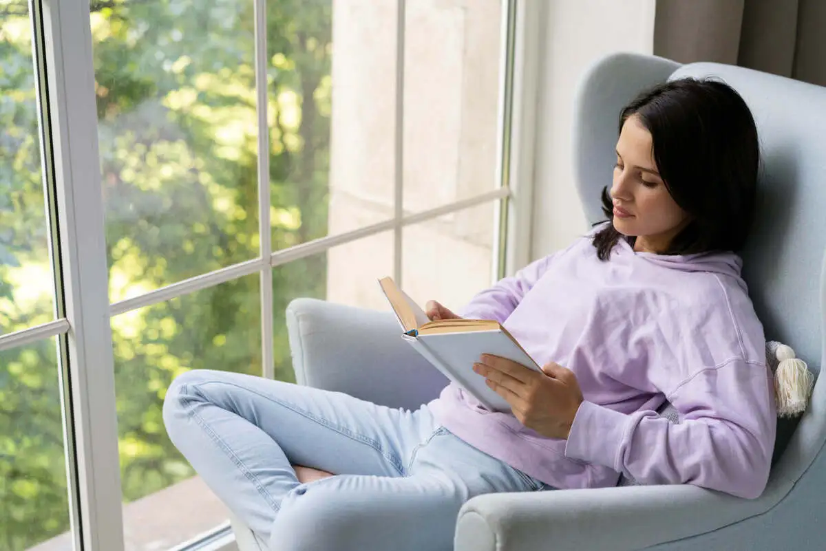 Uma pessoa (homem ou mulher, 30-40 anos) sentada confortavelmente em uma poltrona perto de uma janela com luz natural suave, lendo um livro ou simplesmente olhando para fora com uma expressão calma e satisfeita. O ambiente é limpo, organizado e tranquilo.