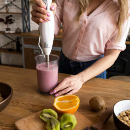 Mulher preparando um smoothie saudável com frutas frescas como kiwi e laranja na cozinha, representando escolhas alimentares que promovem a saúde metabólica e auxiliam no emagrecimento, complementadas pelos produtos da Órion Farmácia de Manipulação