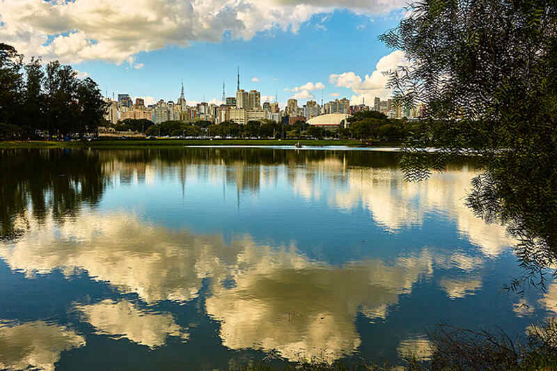 Lago do Parque Ibirapuera em São Paulo, com o reflexo do céu azul e nuvens brancas na água calma, e a silhueta da cidade ao fundo. A imagem transmite uma sensação de tranquilidade, natureza e bem-estar em um ambiente urbano, ideal para promover a qualidade de vida e o equilíbrio que a Órion Farmácia de Manipulação busca oferecer a seus clientes.