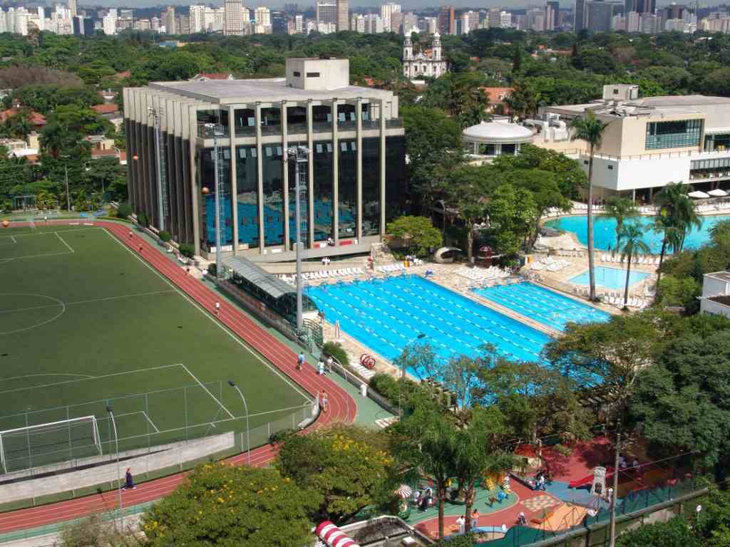 Vista aérea de um clube esportivo e de lazer no Jardim América, São Paulo, com um campo de futebol, pista de corrida, múltiplas piscinas e áreas verdes, emoldurado por edifícios residenciais e comerciais ao fundo. A imagem representa um estilo de vida ativo e saudável, refletindo o compromisso da Órion Farmácia de Manipulação em Jardim América com o bem-estar e a promoção da saúde de sua comunidade.