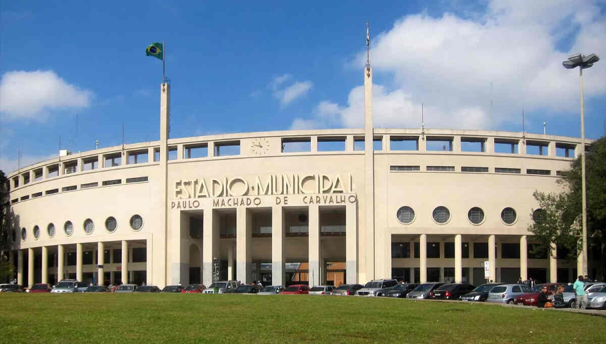 Vista externa do icônico Estádio do Pacaembu, em São Paulo, com sua marcante arquitetura art déco e a rua arborizada em primeiro plano. A imagem representa um dos pontos turísticos e históricos do bairro, destacando a presença da Órion Farmácia de Manipulação no Pacaembu para servir a comunidade local com soluções personalizadas de saúde e bem-estar.