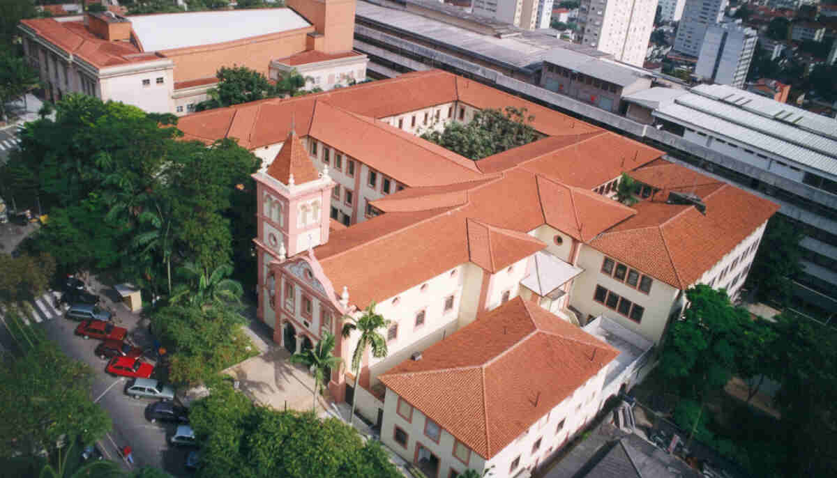 Vista aérea do campus da Pontifícia Universidade Católica (PUC-SP) em Perdizes, São Paulo, com seus edifícios históricos de telhados vermelhos, uma torre de relógio e áreas verdes. A imagem ressalta a localização estratégica da Órion Farmácia de Manipulação em Perdizes, próxima a uma instituição de ensino renomada, reforçando seu compromisso com a ciência, educação e a comunidade local.